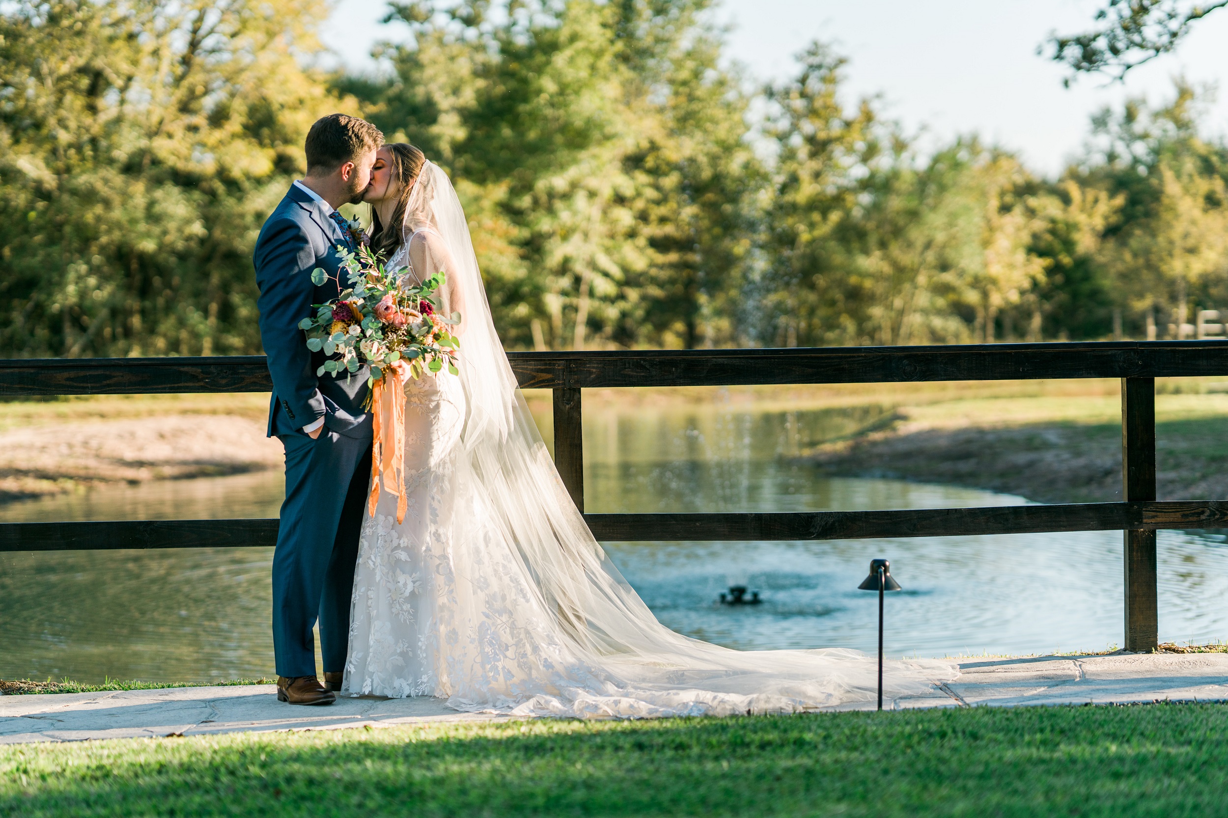 Bride and Groom kissing in front of fenced pond with a water feature as the sun gently lights their faces and background - Houston Wedding Venue
