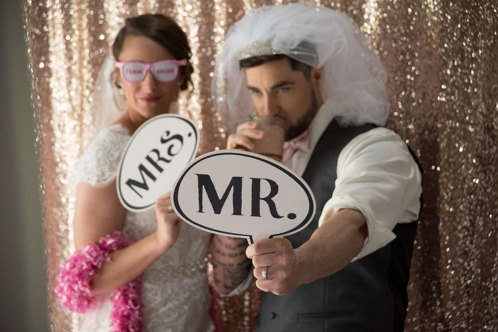 Bride wearing pink shades and pink boa holding a Mrs. sign and Groom holding a Mr. sign standing in front a gold sequins backdrop - Free Wedding Planning Checklist